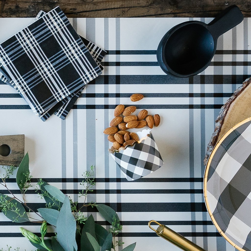 A black and white gingham plaid printed baking cup on a black and white table runner with almonds spilling out. Black and white plaid paper napkins, eucalyptus leaves, a black scooper, and a  black and white plaid, gold rim paper plate.