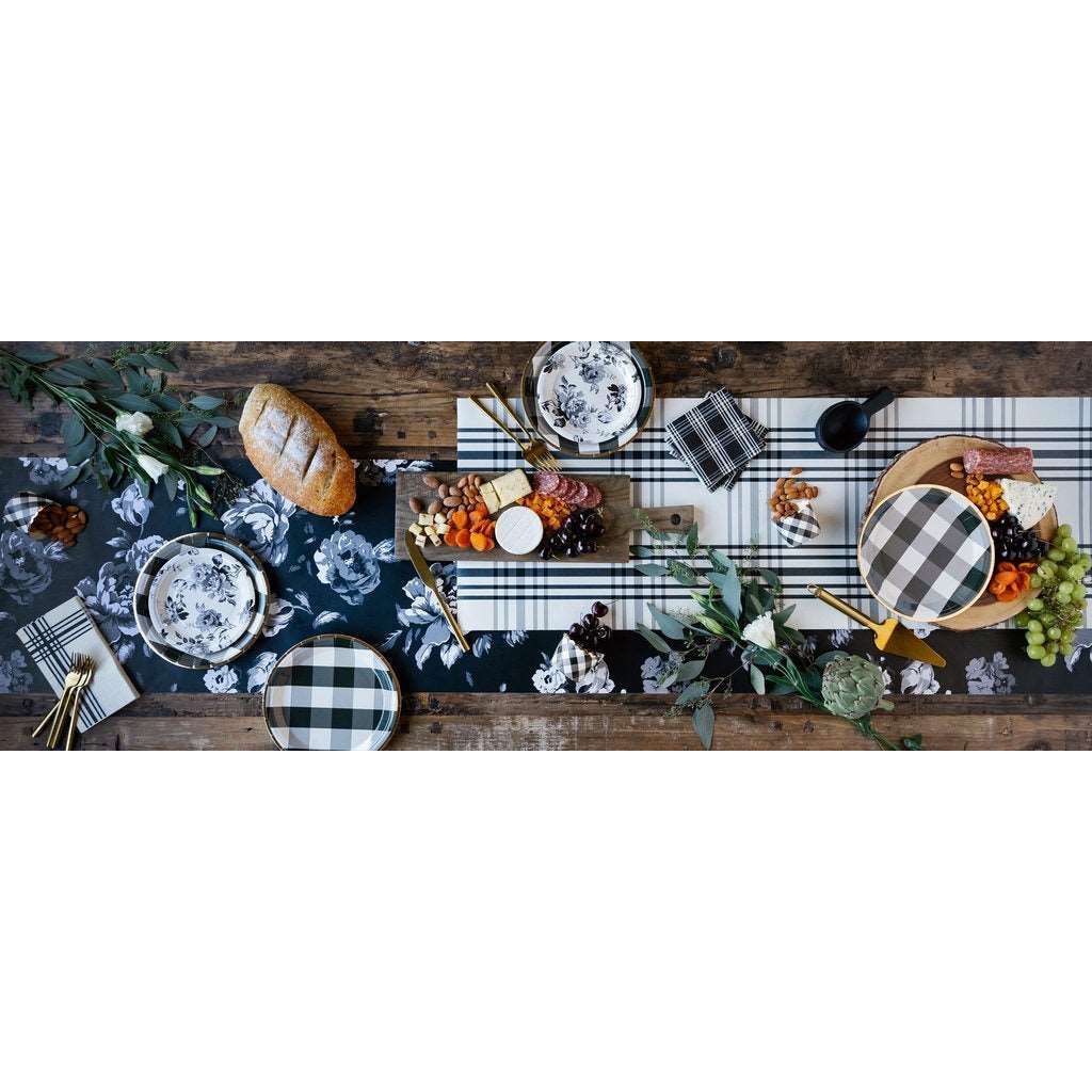 A full table setting, black and white gingham plaid printed baking cups, paper napkins and plates, and gold flatware on a black and white table runner on a rustic wooden table filled with breads , flowers, and cheese, nut, and meat platters.