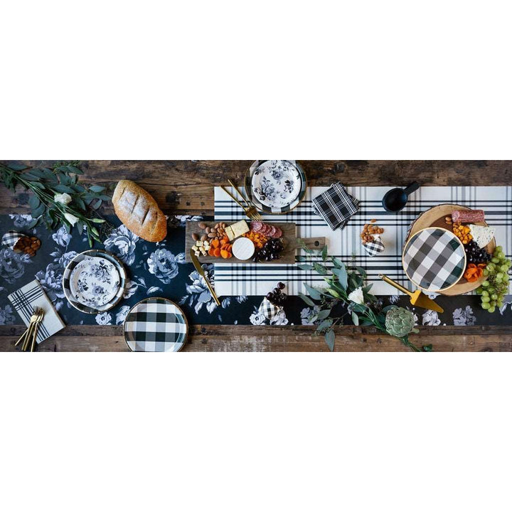 A full table setting, black and white gingham plaid printed baking cups, paper napkins and plates, and gold flatware on a black and white table runner on a rustic wooden table filled with breads , flowers, and cheese, nut, and meat platters.