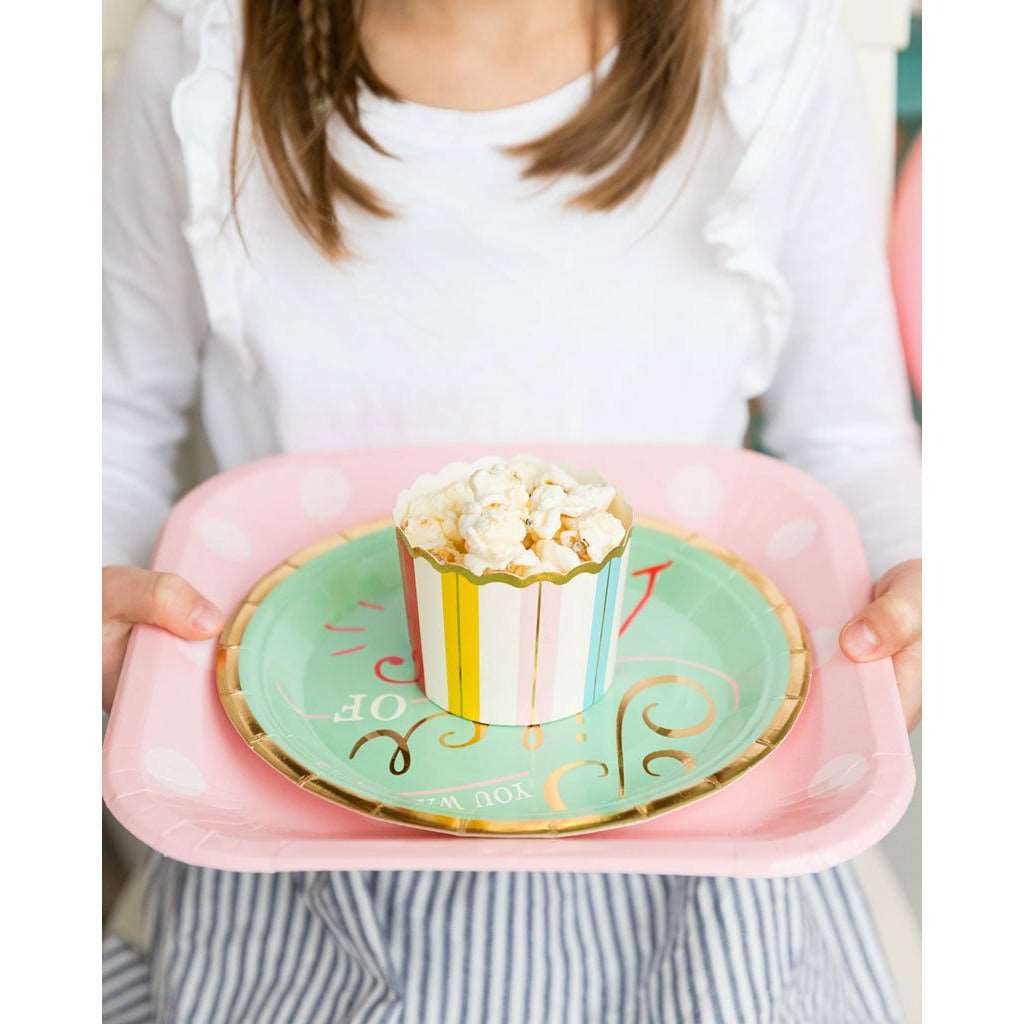 a woman holding a pink paper charger plate, with a mint green paper dinner plate on top with a rainbow striped treat cup filled with popcorn on top.