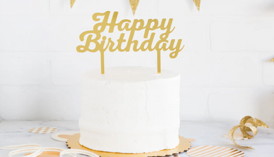 White cake with gold 'Happy Birthday' topper on a white and gold decorated background