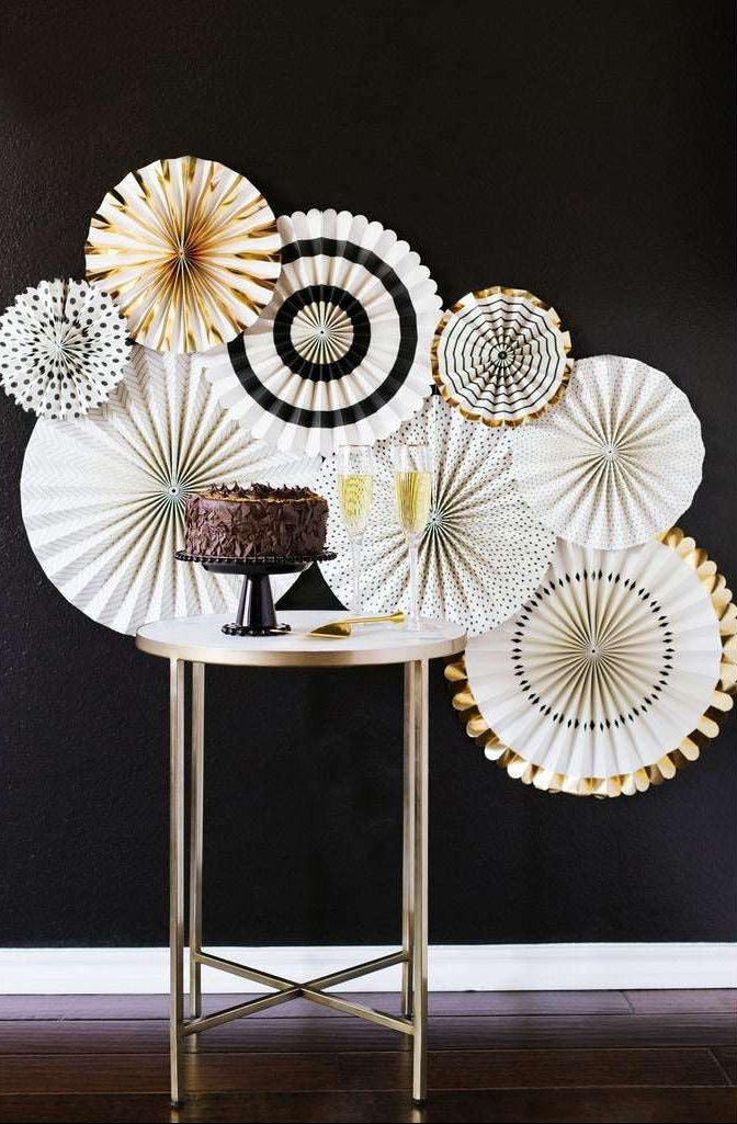 A set of elegant paper fans with black, cream, and gold patterns in varying sizes and print patterns against a black wall with a chocolate cake on a black cake stand and champagne glasses on a gold marble top table.