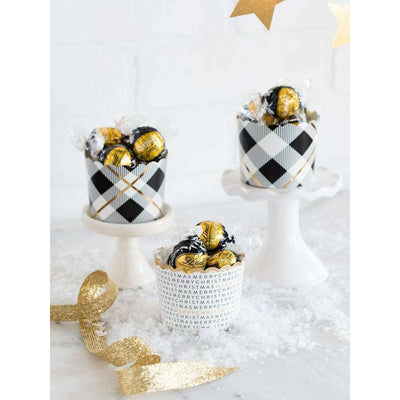 Two black, white and gold printed plaid patterned cupcake-treat cups each on a white cupcake stand and one white cupcake-treat cup with the words "merry christmas" repeated in multiple lines around the cup in black and gold print, displayed on a white surface with Christmas-themed decorations in the background.