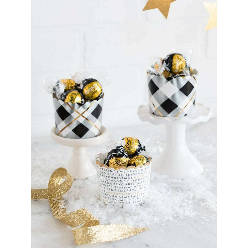 Two black, white and gold printed plaid patterned cupcake-treat cups each on a white cupcake stand and one white cupcake-treat cup with the words "merry christmas" repeated in multiple lines around the cup in black and gold print, displayed on a white surface with Christmas-themed decorations in the background.