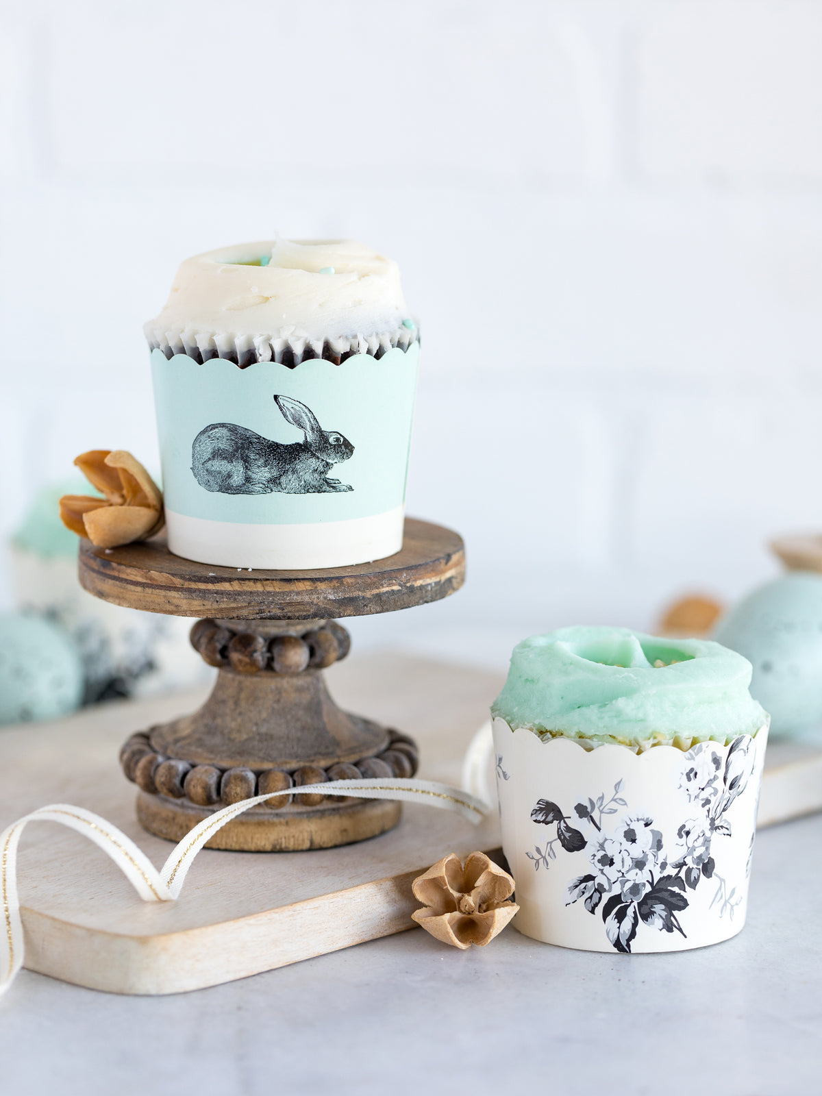 Tiffany blue and cream cupcake cup with a black rabbit printed on it with a vanilla frosted cupcake in it on a wooden cupcake stand on a wood cutting board, next to the cutting board a cream cupcake cup with black floral print and a tiffany blue frosted cupcake in it