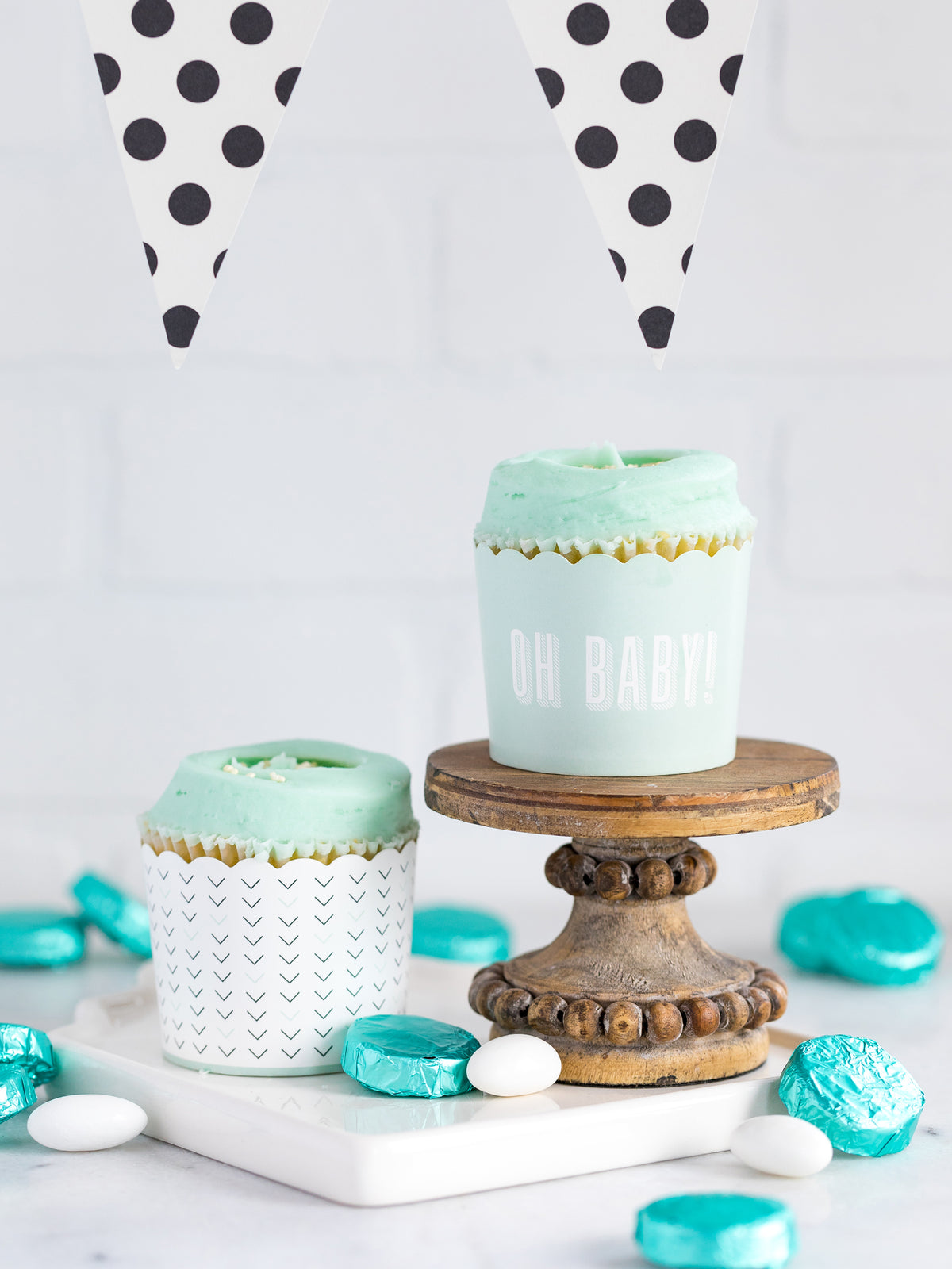 A set of baking cups with one design featuring 'Oh Baby' text in cream print on a mint green background, and another with a black and mint green all-over v print. The cups are displayed on a wooden stand and a white surface with decorative polka dots in the background.
