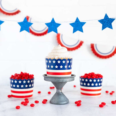 White background, white table with red jellybeans on it. blue star banner and a red, white, and blue bunting banner both hanging in the background. 3 baking cups with red and white stripes from bottom to the center of cup and blue stripe with white stars from center to the top of cup.