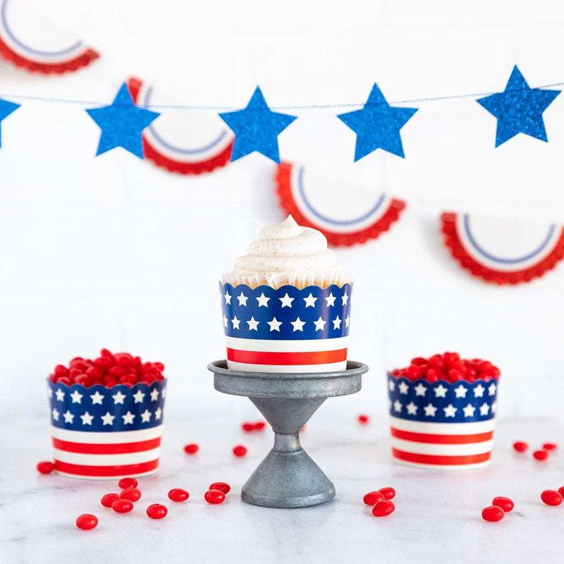 White background, white table with red jellybeans on it. blue star banner and a red, white, and blue bunting banner both hanging in the background. 3 baking cups with red and white stripes from bottom to the center of cup and blue stripe with white stars from center to the top of cup.
