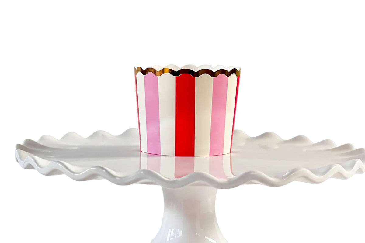 Red, pink, and white striped cupcake cup with gold foil scalloped rim on a white cake stand with a kitchen background