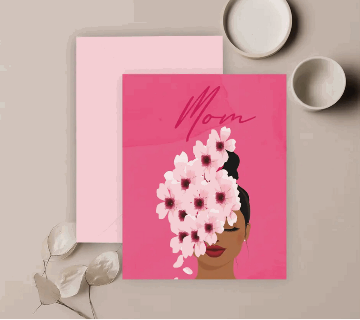 Hot pink greeting card with an illustration of a woman with white and pink flowers cascading down from her bun to her face and the word "Mom" in deep pink written above her head, a light pink envelope. All against a neutral background with decorative objects