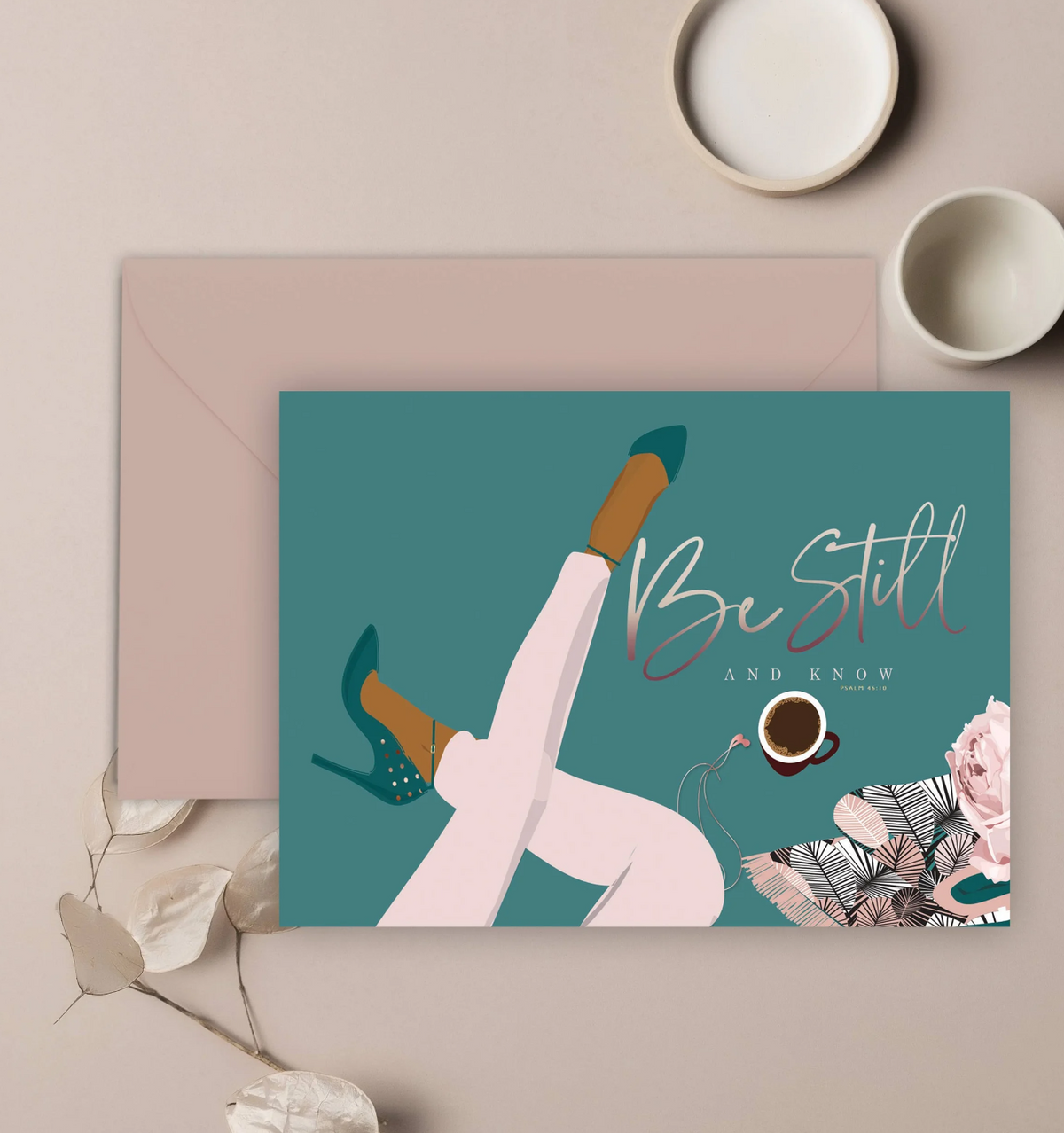 A teal colored greeting card with an illustration of a woman's crossed legs and heeled shoes, a cup of coffee, ear pods, a floral and feather sketch in the bottom right corner, and the words be still in script, "and know psalms 46:10" in plain text, and a mauve colored envelope, displayed on a neutral background.