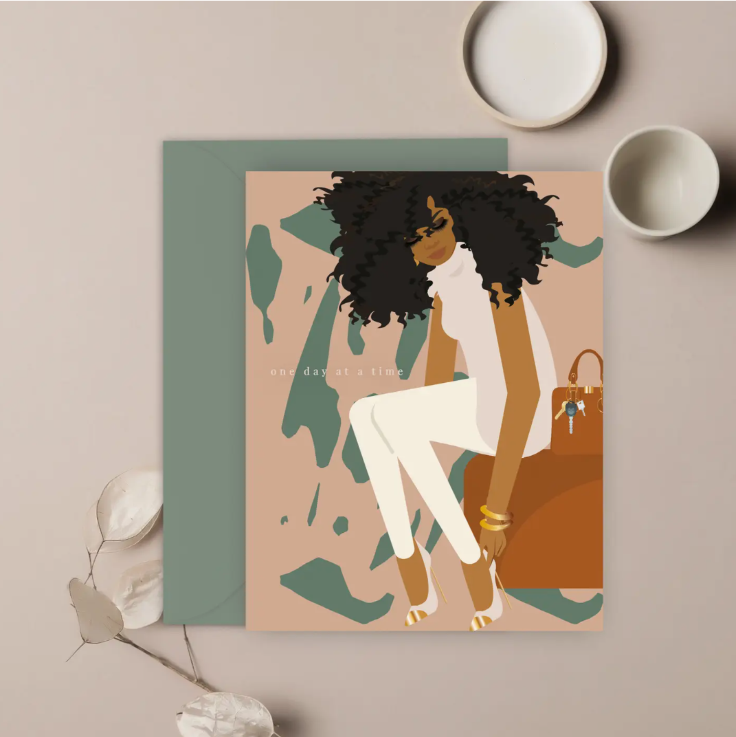 A greeting card featuring an illustration of a woman with curly hair sitting next to a handbag, with abstract shapes in the background. The card is placed next to a beige envelope and some decorative elements on a neutral-toned surface.