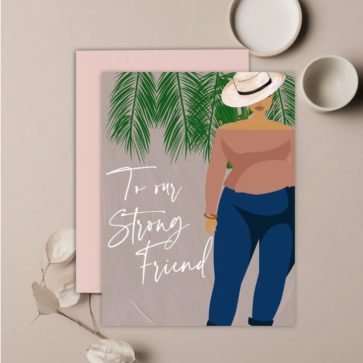 A tauped colored greeting card with white text that reads "To Our Strong Friend" with an illustration of a woman wearing a white fedora hat, a boat-neck top, and blue jeans, standing among tropical leaves. The card is placed on a neutral background.