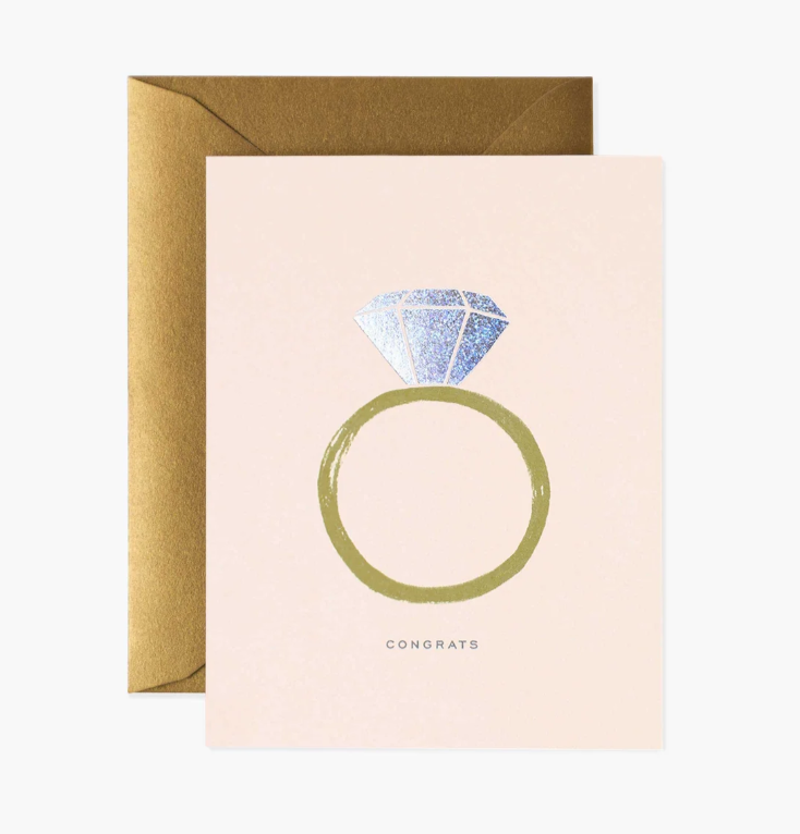 Blush pink greeting card with a gold band diamond ring illustration with iridescent diamond, and the word "Congrats" in grey print below it, with a metallic bronze envelope