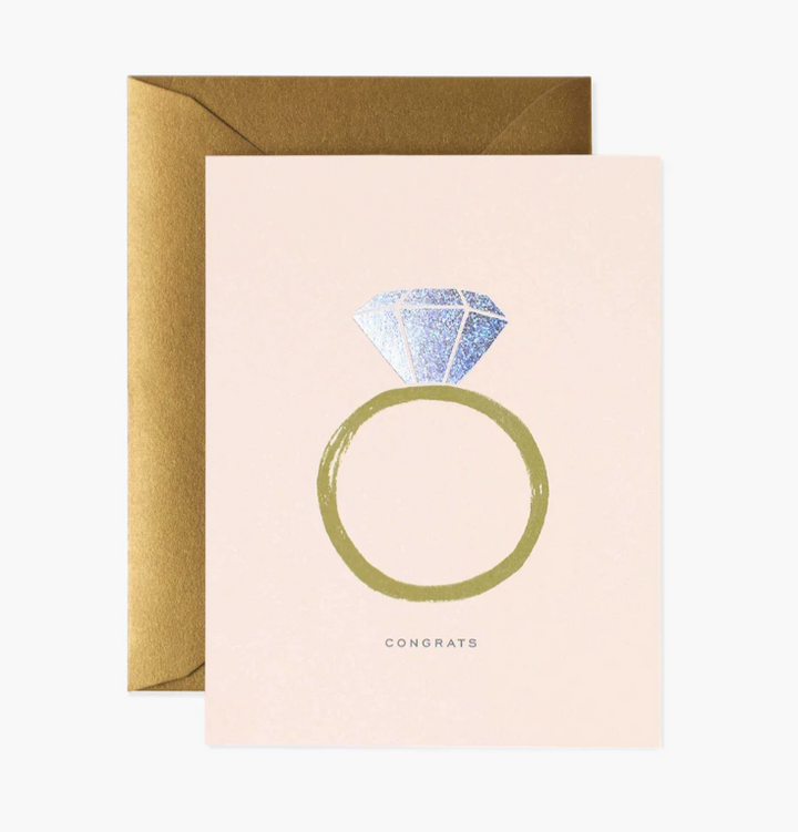 Blush pink greeting card with a gold band diamond ring illustration with iridescent diamond, and the word "Congrats" in grey print below it, with a metallic bronze envelope