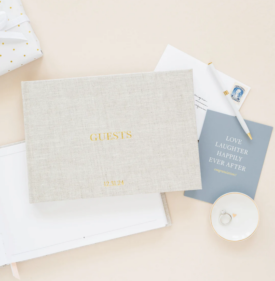 Natural color fabric bookcloth covered guest book with the word guest in gold foil print on a pink surface with stationery items underneath it.