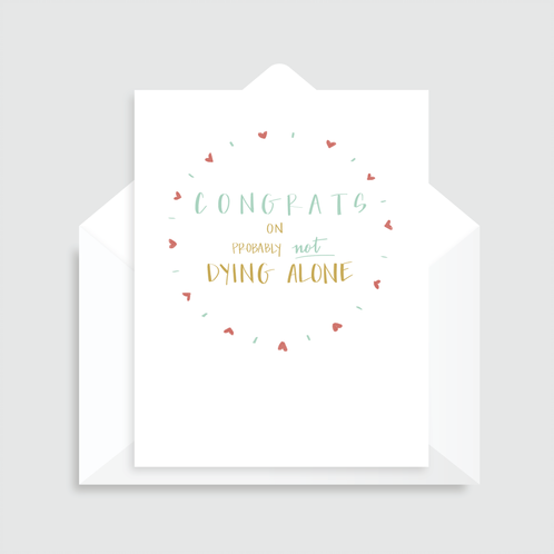 White greeting card with illustrated mini red hearts creating a circle with text inside that reads "Congrats on probably not dying alone" paired with a white envelope
