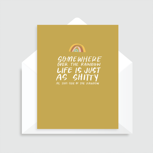 Mustard greeting card with an illustrated rainbow at the top and white text that reads "Somewhere over the rainbow life is just as shitty as this side of the rainbow". Paired with a white envelope.