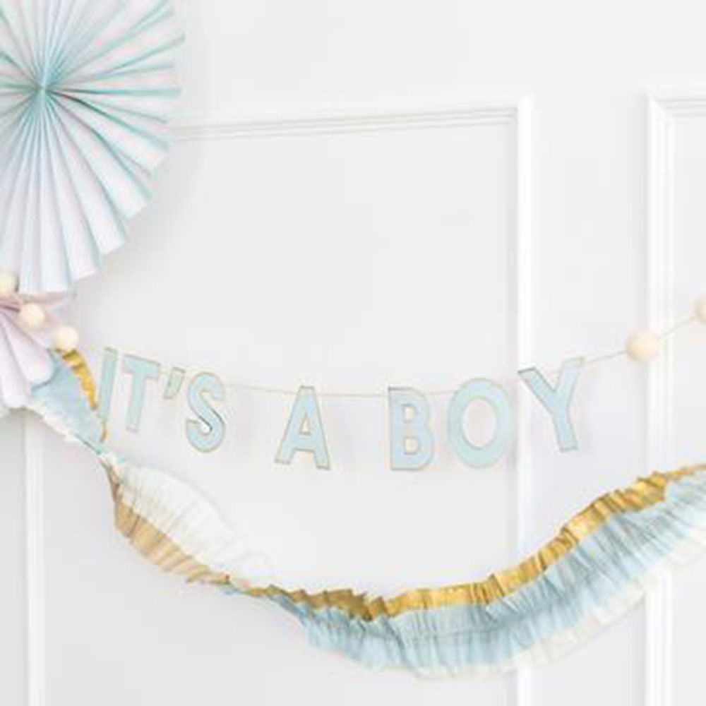 It's A Boy Letter Banner with pom poms hanging on a wall with blue, gold, and white tissue banner and a blue party fan against a white background