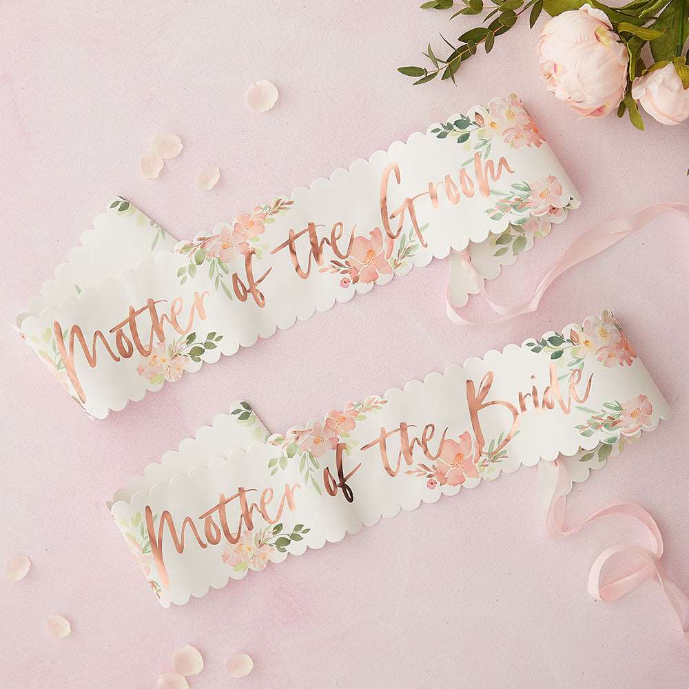 Two paper sashes with floral patterns and 'Mother of the Bride and Mother of the Groom' printed in rose gold foil on a pink background, surrounded by pink peonies and greenery