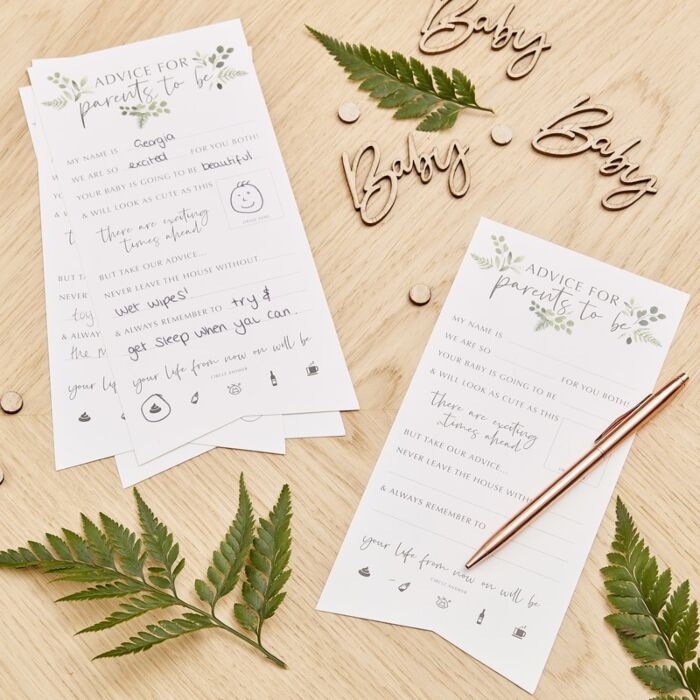 A set of white advice cards for new parents with a white background and green botanical illustrations on a wooden table surrounded by wooden letters spelling the word baby and fern stems.