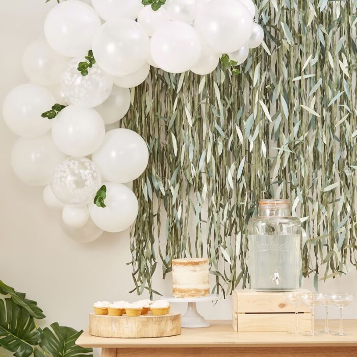 A combination of solid white and clear white confetti-filled balloons with green eucalyptus sprigs against a grass wall above a wooden table with cupcakes, a beverage dispenser, wine glasses, and a cake.