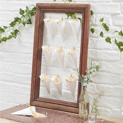 A display of white scalloped-edge confetti cones arranged in a wooden frame on a sequins runner lined table, with a decorative wall in the background. Cones are filled with rose petals or rice, and there is one cone of confetti on the table in front of the frame.