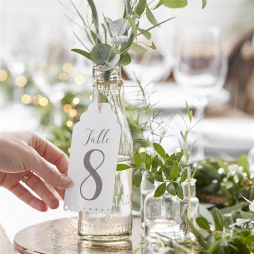 white table number card with scalled eyelet detail at bottom edge, grey printed table numbers hanging on a bottle filled with water filled with eucalytpus stems.