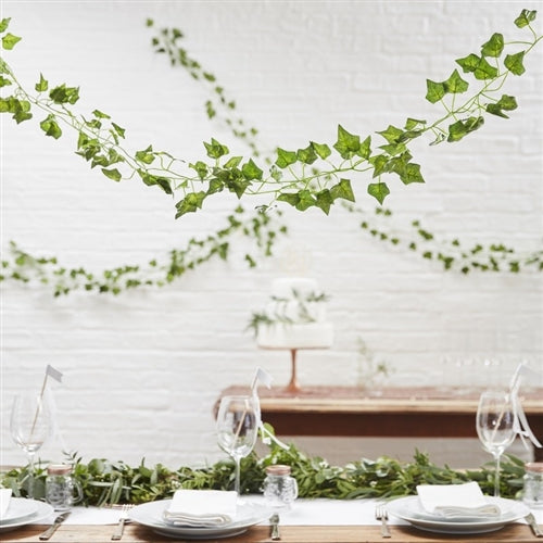 Numerous ivy garlands swagged from the ceiling  over a wooden table with a dinner table setting and a wedding cake in the background