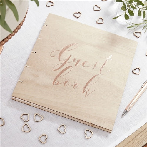 Wooden guest book with twine binding and a rose gold foil printed guest book on the front on a table with heart shaped wooden confetti around it, a pen, and foliage.