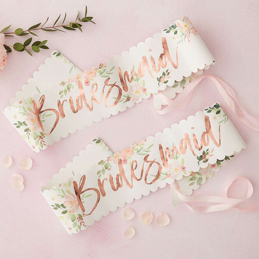 Two paper sashes with floral patterns and 'Bridesmaid' printed in rose gold foil, tied with pink ribbons on a pink background surrounded by pink rose petals and greenery