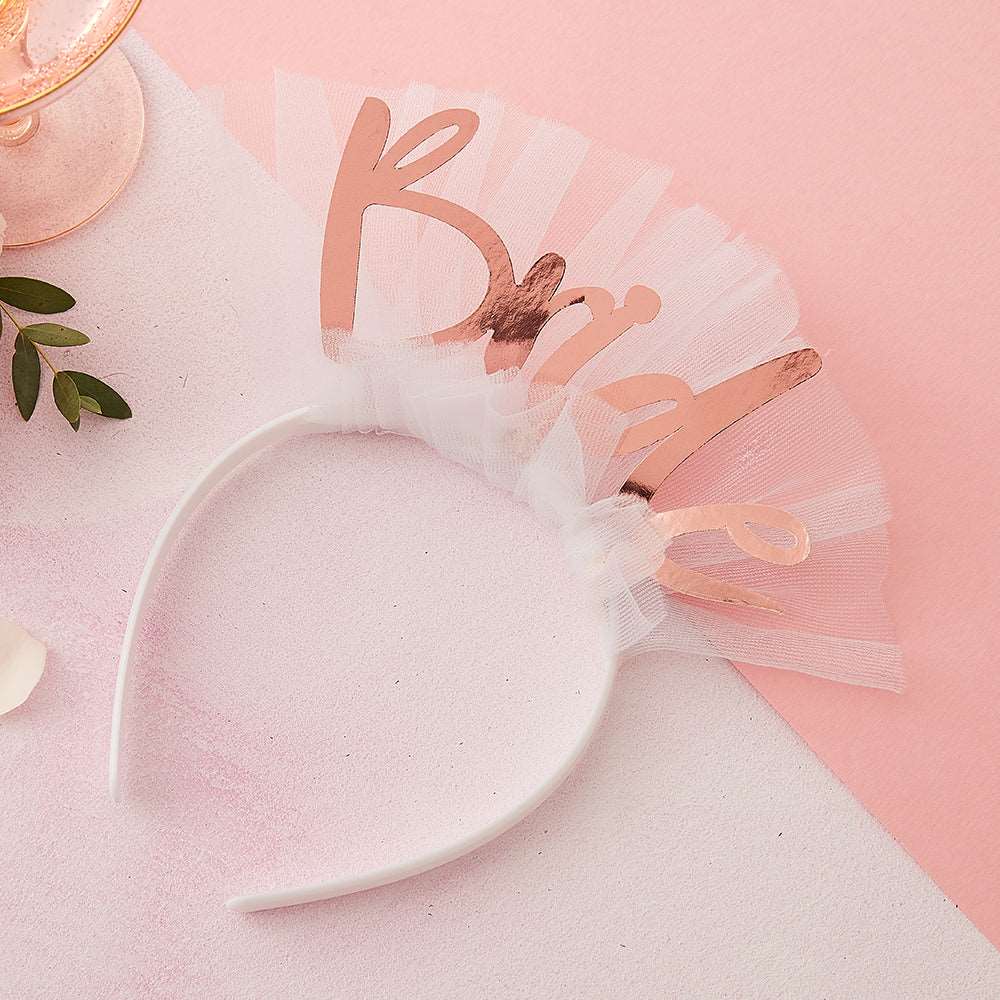 White headband wrapped in white tulle with bride in rose gold individual letters on top, lying on a white and pink textured background next to a wine glass and foliage.