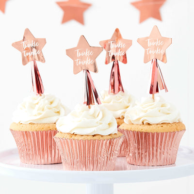 Set of 4 rose gold foil, star-shaped cupcake toppers with tassels and phrase “Twinkle Twinkle” in script on the star placed on top of cupcakes on a white table.