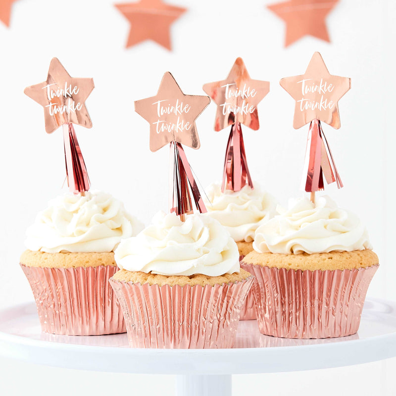 Set of 4 rose gold foil, star-shaped cupcake toppers with tassels and phrase “Twinkle Twinkle” in script on the star placed on top of cupcakes on a white table.