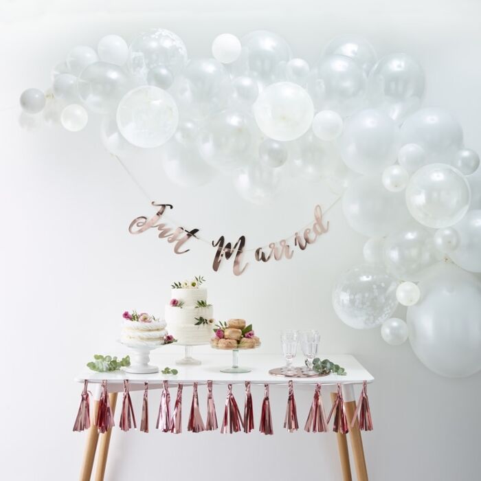A white balloon arch arranged with various-sized and finished balloons above a dessert table and a just married  letter banner in rose gold color