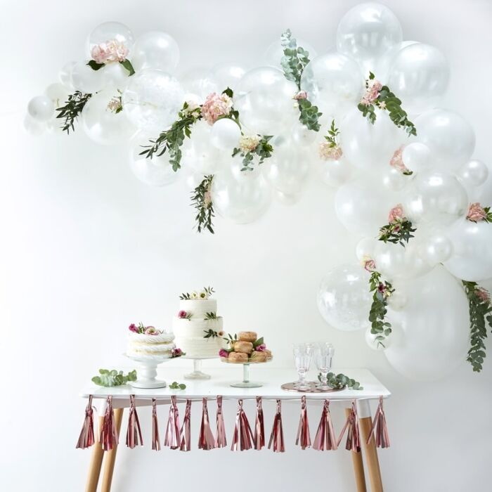 A white balloon arch arranged with various-sized and finished balloons above a dessert table and a rose gold tassel banner on the table edge