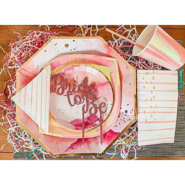 Peachy-pink iridescent paper cup and matching straw laying on a table next to a tonal pink and white splash printed gold rim paper dinner and salad plate, gold dessert plate with pink, white and gold striped napkins on a wooden table