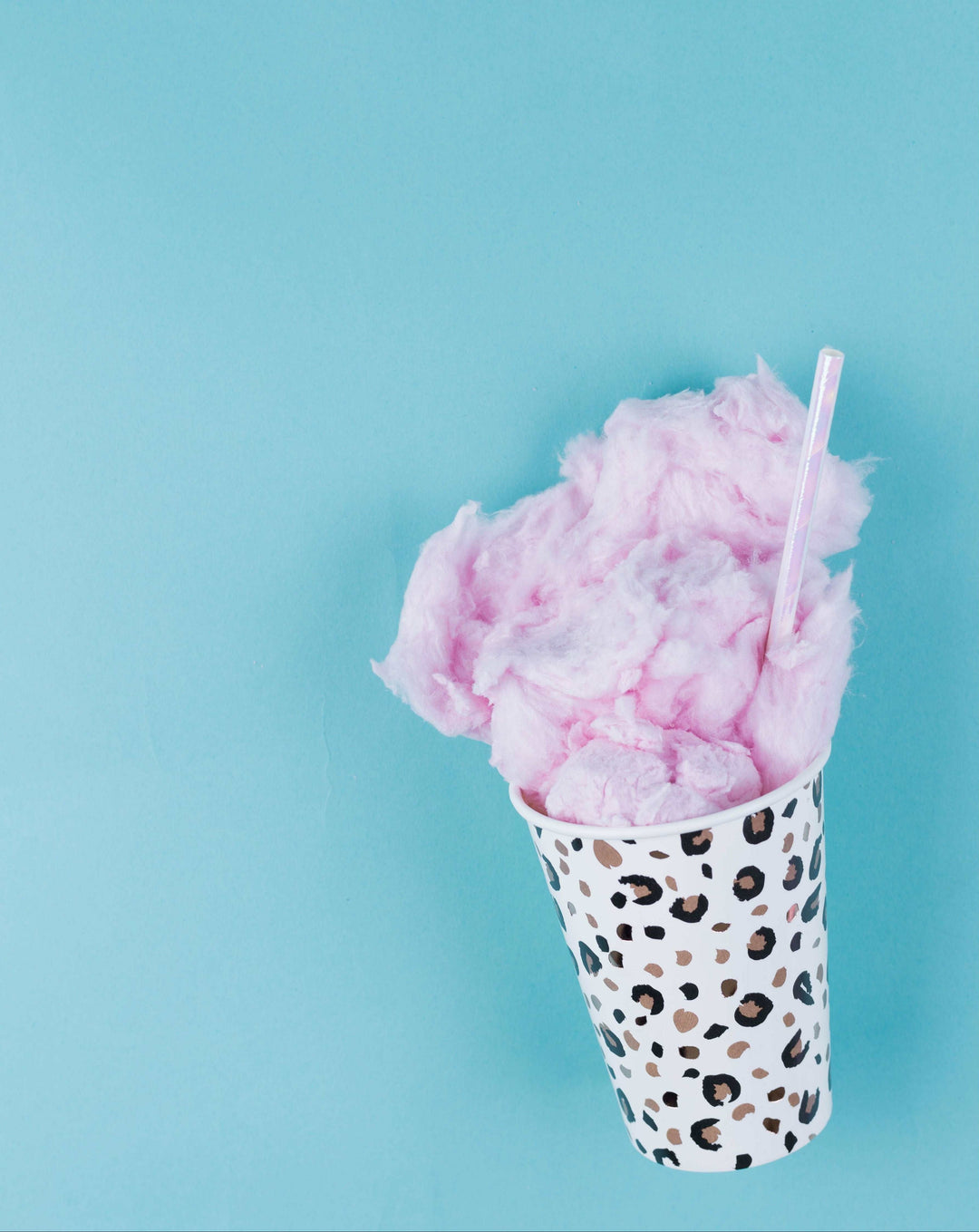 Leopard printed paper cup with cotton candy coming out of it and a pink iridescent straw on a teal blue background