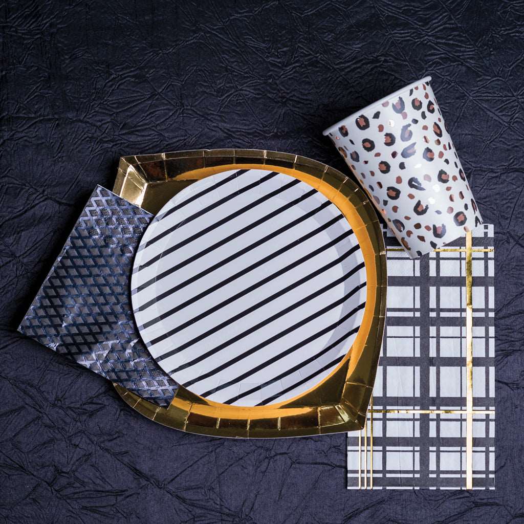 Table setting of gold foil party plate, white and black striped party plate, animal print cup, white, black, and gold foil napkin, and a black textured napkin.