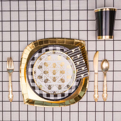 gold charger plate topped with black and white checker print dinner plate, topped with a white dessert plate with allover gold bumble bee print, black and white striped napkin with allover gold bumble bee print, gold flatware set, and a black cup with gold borders on top of a white and black checker print tablecloth.