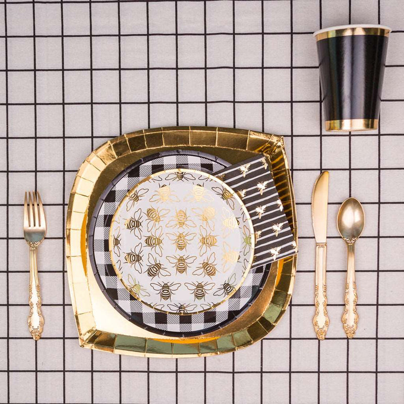 gold charger plate topped with black and white checker print dinner plate, topped with a white dessert plate with allover gold bumble bee print, black and white striped napkin with allover gold bumble bee print, gold flatware set, and a black cup with gold borders on top of a white and black checker print tablecloth.