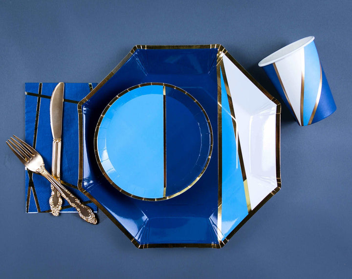 overhead view of a dinnerware set, a navy, sky blue, white, and gold printed charger plate, sky blue and navy with gold foil trim printed dessert plate, navy, sky blue, white, and gold foil swirl print paper cup, and gold fork and knife on a jewel tone navy nakin with gold foil print pattern against a navy background