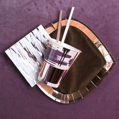 wine colored suede background, with rose gold foil paper dinner plate topped with a white with rose gold zebra print napkin, a paper cup with a brushstroke pattern in mauve, taupe, and tan colors with rose gold accents, and a rose gold drinking straw in it