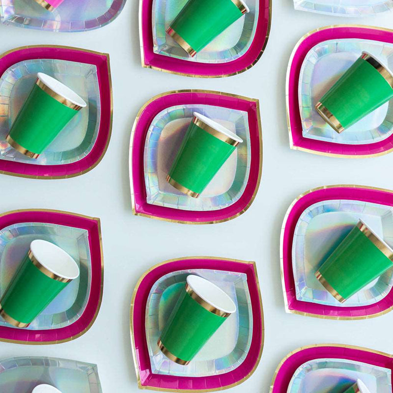 a group of hot pink charger plates with gold rim, topped with iridescent silver dinner plates, topped with green paper cup with gold borders on a white background