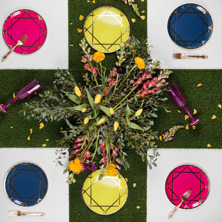 Turf-style tablecloth topped with gemstone-inspired paper plate set, 2 sapphire blue with gold accent dinner plates, 2 fuchsias with gold accent dinner plates, and 2 yellow with gold accent dinner plates, gold forks and purple wine glasses with a floral arrangement centerpiece