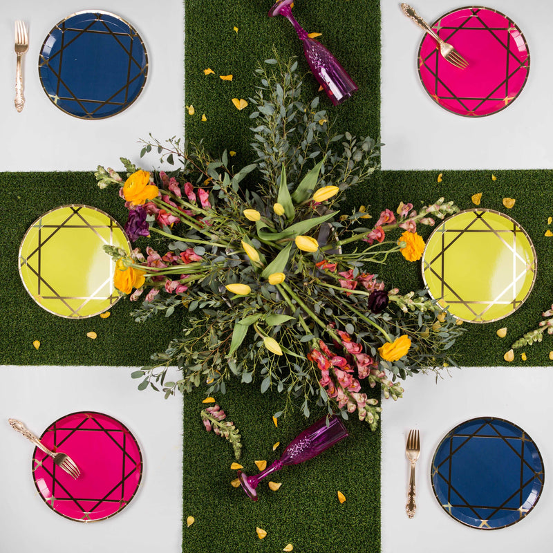 Turf-style tablecloth topped with gemstone-inspired paper plate set, 2 sapphire blue with gold accent dinner plates, 2 fuchsia with gold accent dinner plates, and 2 yellow with gold accent dinner plates, gold forks and purple wine glasses with a floral arrangement centerpience