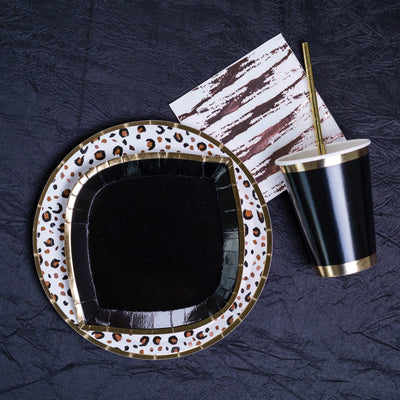 Black fabric background with an animal printed gold rim paper plate, topped with a black with gold rim paper plate, next to a black and gold trimmed paper cup with a gold paper straw in it laying down, and a white paper napkin with a rose gold zebra stripe print