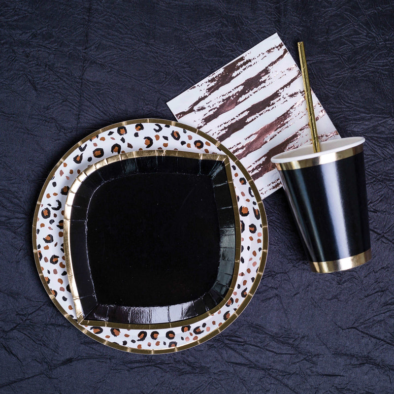 Black fabric background with an animal printed gold rim paper plate, topped with a black with gold rim paper plate, next to a black and gold trimmed paper cup with a gold paper straw in it laying down, and a white paper napkin with a rose gold zebra stripe print