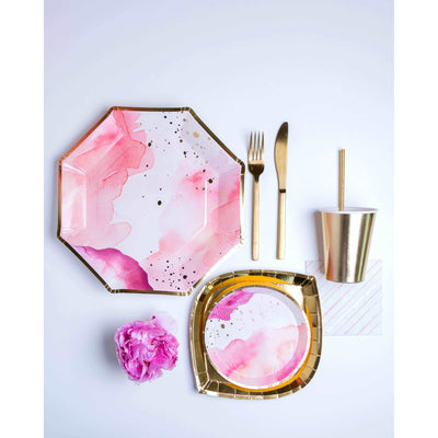 Top view of tonal pink and white splatter print pattern, gold rimmed dinner and dessert paper plate, gold foil salad paper plate, gold foil paper cup and straw, a pink and gold diagonal striped cocktail paper napkin, gold fork and knife, and a pink peony flower on a white background
