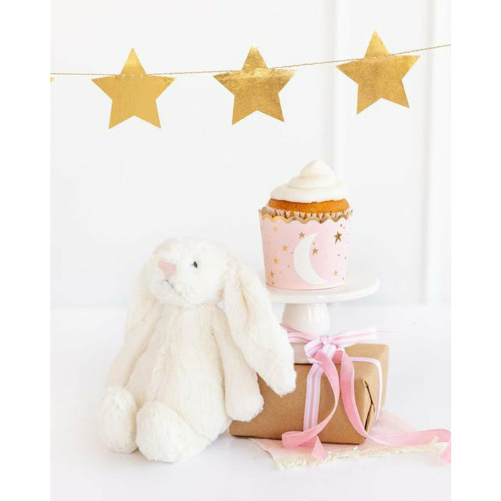 Pink cupcake cup with a cupcake topped with vanilla frosting on top of a box. gold star banner above and a stuffed rabbit toy and pink ribbon